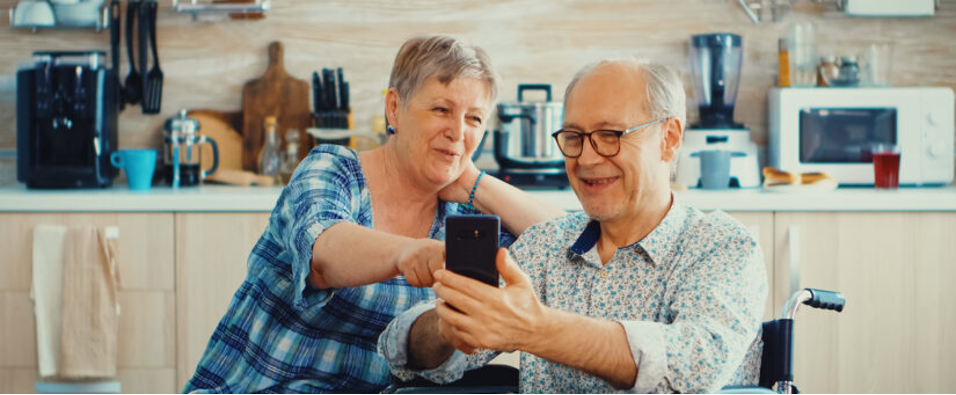 Elderly woman and elderly man smiling and looking at a cell phone screen, in the background, there is a kitchen. The woman is white, with short gray hair, wears a blue plaid blouse, is sitting and pointing at her cell phone screen. The man is white, wears glasses, has short white hair, wears a white and blue shirt, is sitting in a wheelchair and holding his cell phone in front of his chest with both hands.