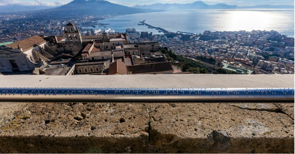 In the foreground, there is a silver handrail with Braille engraving all over it. In the background, there are several old buildings, the coast of a sea reflecting the sunlight with mountains in the far back.